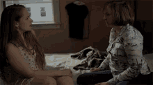 two women are sitting on a bed talking to each other and looking at each other .