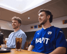 two men are sitting at a table with one wearing a shirt that says napis
