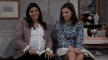 a pregnant woman sits next to another woman in front of a framed picture that says ' double trouble ' on it