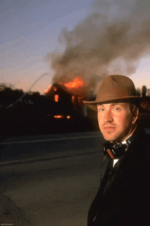 a man wearing a cowboy hat and headphones stands in front of a burning house