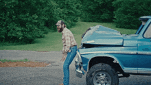 a man in a plaid shirt is standing next to a blue truck that has been damaged