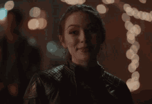 a woman in a black leather jacket smiles in front of a string of lights