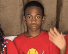 a young boy wearing a red t-shirt with a softball on it is waving his hand .