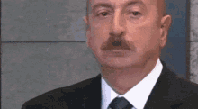 a man with a mustache wearing a suit and tie looks at the camera