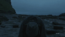 a man in a hooded cape stands on a rocky beach with the letters rmjzl on the bottom right