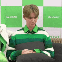a man in a green and white striped shirt sits on a couch with iq.com written on the wall behind him