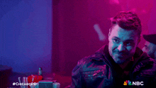 a man in a leather jacket stands in a dark room with a nbc logo on the bottom right