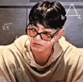 a young man wearing glasses is sitting in front of a chalkboard with math equations on it .