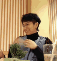 a woman wearing glasses and a black turtleneck is smiling while holding a plant