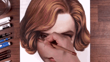 a person is drawing a woman 's face with a pencil on a wooden table with pens and markers