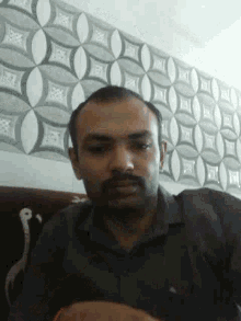 a man with a beard is sitting in front of a wall with a pattern on it