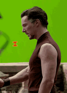a man in a brown tank top stands in front of a green screen that says i