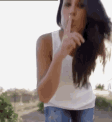 a woman in a white tank top and blue jeans is making a shhh gesture with her finger .