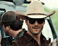 a man wearing a cowboy hat and sunglasses is smiling in front of a car .