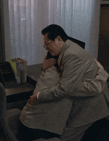a man in a suit and glasses is hugging another man in a chair