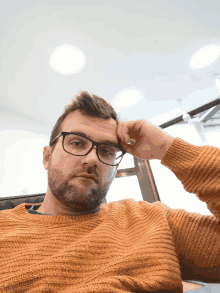 a man wearing glasses and a yellow sweater holds his hand to his face