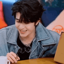 a young man in a denim jacket is sitting at a table with a cup of coffee and a laptop .