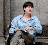 a man wearing glasses sits on a bench holding a notebook and a pen