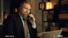 a man in a suit and tie is talking on a phone with the nbc logo behind him