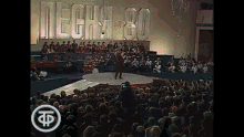 a man stands on a stage in front of a crowd with a sign that says mecha-80