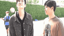two young men are standing next to each other in front of a hedge and looking up .