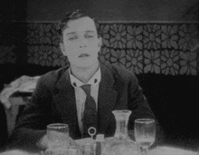 a man in a suit and tie sits at a table with a decanter and glasses