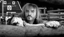 a black and white photo of a man with long blonde hair and a beard leaning on a log .