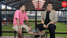 two men sit at a table in front of a fence that says poros malang