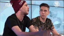 two young men are sitting next to each other and one has a supreme hat on