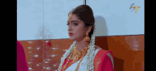 a woman in a pink and white saree and gold jewelry is standing in front of a wall .