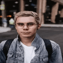 a young man wearing glasses and a denim jacket is walking down a street .