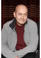 a bald man wearing a grey jacket and a red shirt looks at the camera