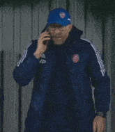 a man wearing a blue jacket and a blue hat with arsenal on it talks on a cell phone