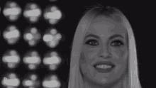 a black and white photo of a woman with long blonde hair smiling in front of a wall of lights .