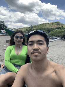 a man and a woman pose for a picture on a beach with a boat in the background
