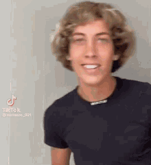 a young man with curly blonde hair is wearing a black shirt and smiling .