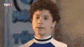 a young boy with curly hair is standing in front of a sign that says trt 1