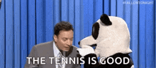 a man in a suit and tie is talking to a stuffed panda bear who says `` the tennis is good '' .
