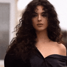 a woman with long curly hair is wearing a black off the shoulder top and looking at the camera .