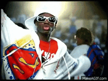a man wearing a hat , sunglasses and a superman shirt is standing in a crowd .