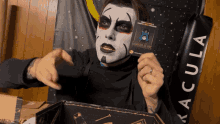 a man with white face paint holds a card that says dracula