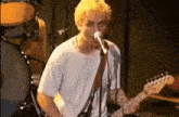 a man with yellow hair is singing into a microphone while playing a guitar on stage .