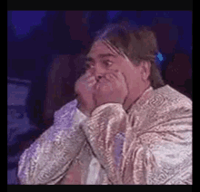 a man with long hair is covering his mouth with his hands while wearing a gold jacket .