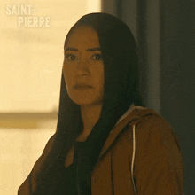 a woman with long black hair is wearing a brown hoodie and looking at the camera .