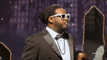a man wearing sunglasses and a beanie stands in front of a screen that says adult swim.com