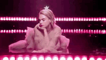 a woman in a pink dress with a star on her head is sitting in front of a row of pink lights .