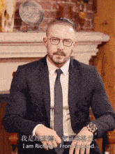 a man in a suit and tie is sitting in front of a fireplace and says " i am richard from england "