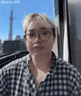 a woman wearing glasses and a plaid shirt is standing in front of a window with a skyscraper in the background