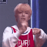 a young man with blonde hair is wearing a red basketball jersey and giving a thumbs up .