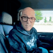 a bald man wearing glasses and a tie dye hoodie is sitting in the back seat of a car